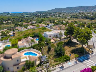 Vista aérea de una parcela residencial llana con árboles maduros, junto a una carretera con paso de cebra. Ofrece vistas lejanas al mar y a las verdes colinas circundantes.