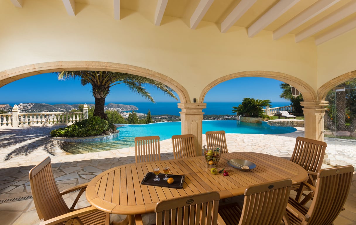 Amplia naya cubierta con mesa de comedor de madera y sillas, ofreciendo vistas panorámicas al mar Mediterráneo y la costa a través de elegantes arcos de piedra y una piscina infinity con palmeras.