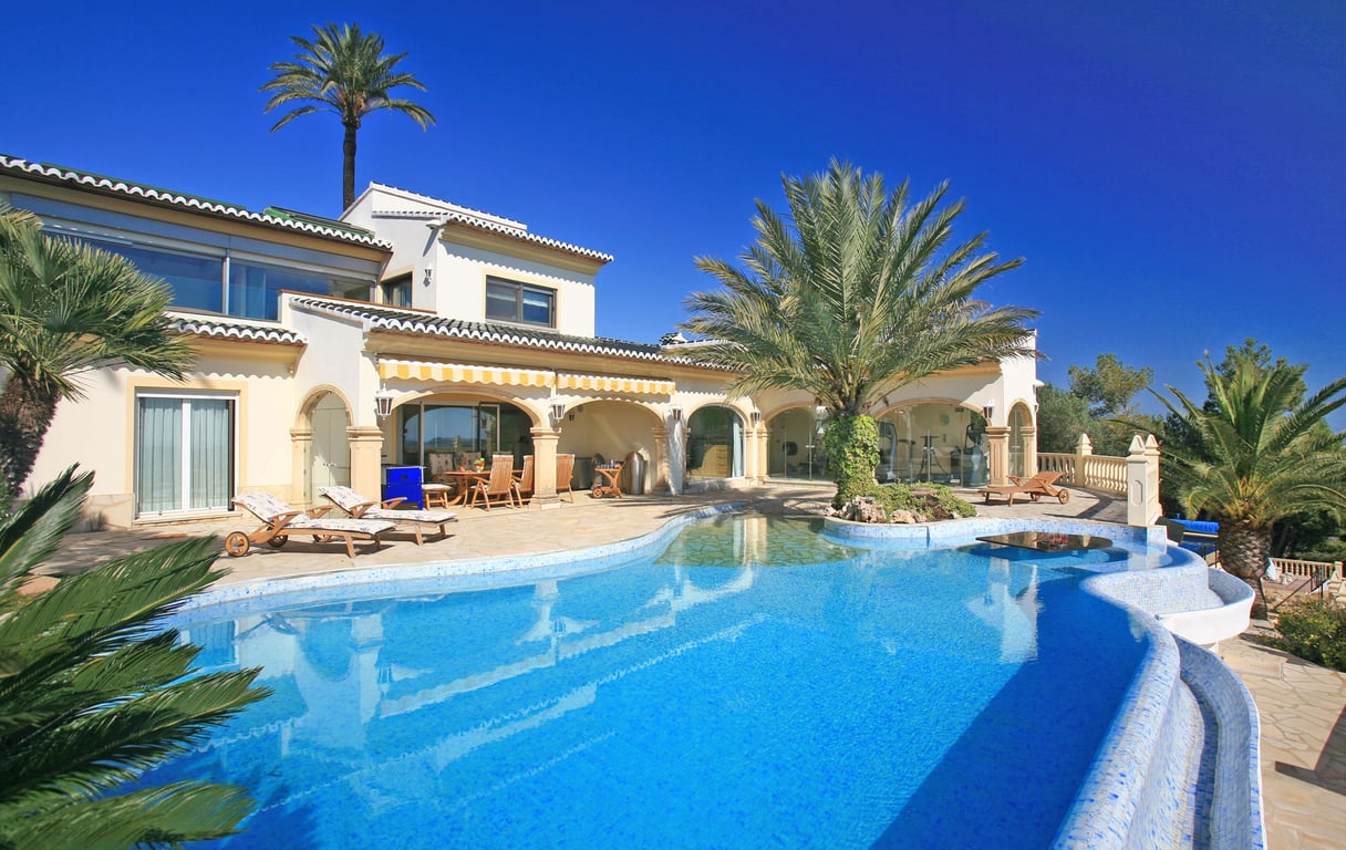 Amplia piscina de mosaico azul con entrada tipo playa, isla de palmeras y mesa acuática. La villa ofrece una naya cubierta y un gimnasio acristalado.