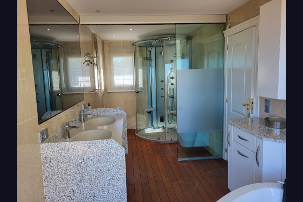 Amplio baño con doble lavabo de diseño en guijarro, ducha circular de hidromasaje y suelos de madera. Incluye espejo grande y ventanas con persianas.