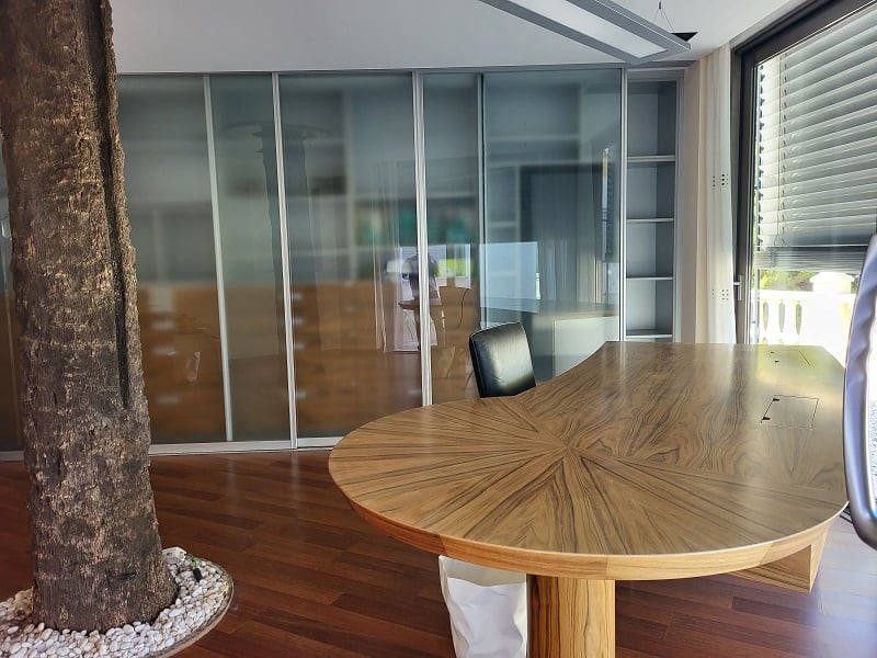 Amplio despacho privado con un escritorio de madera de diseño único, puertas correderas de cristal esmerilado y un árbol interior, complementado por suelos de madera y luz natural.