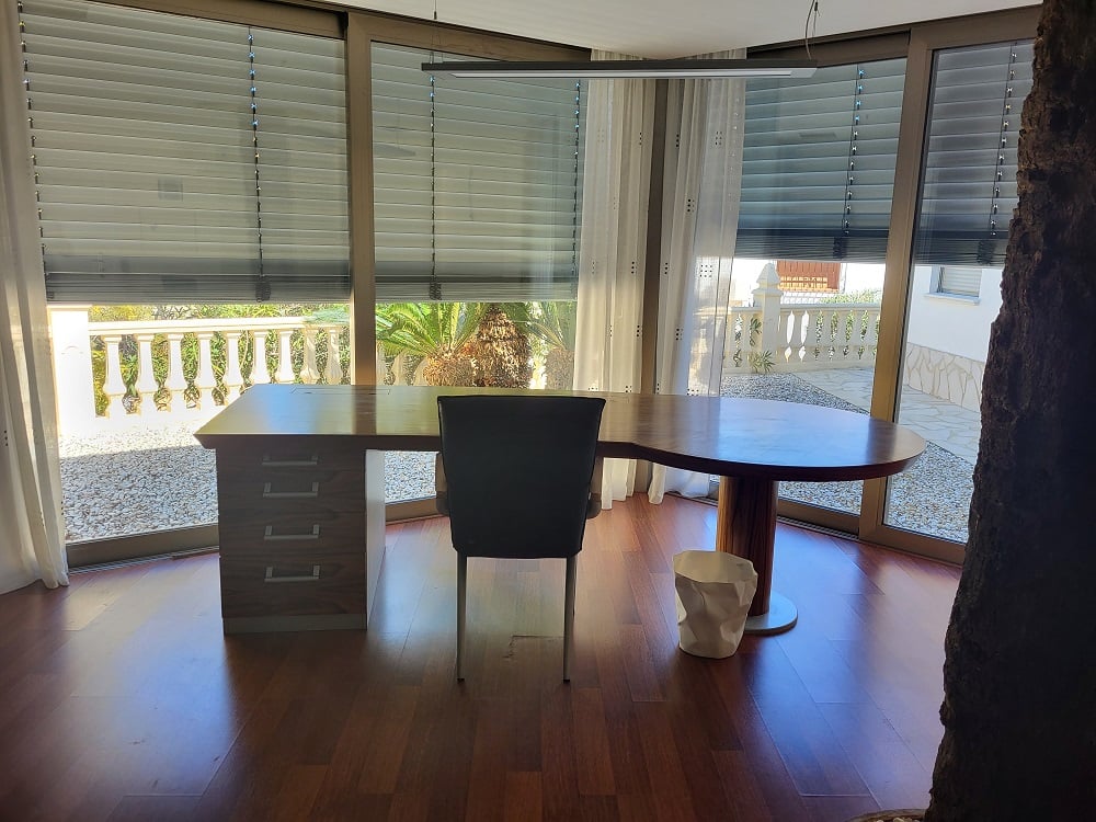 Amplio despacho privado con un escritorio de madera de diseño y grandes ventanales que ofrecen vistas a un patio ajardinado. El suelo de parquet oscuro y la iluminación moderna completan el espacio.