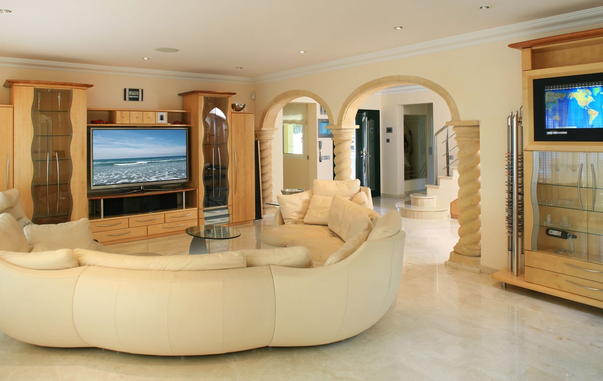 Amplio salón con sofá curvo color crema, suelos de mármol claro y muebles de entretenimiento de madera clara a medida. Arcos de piedra con columnas decoradas conectan con la entrada.