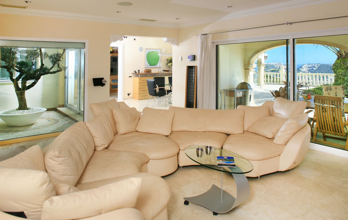 Amplio salón con sofá seccional claro, mesa de centro de cristal y suelos de mármol. Grandes ventanales conectan con una terraza con vistas al mar y un patio interior con olivo.