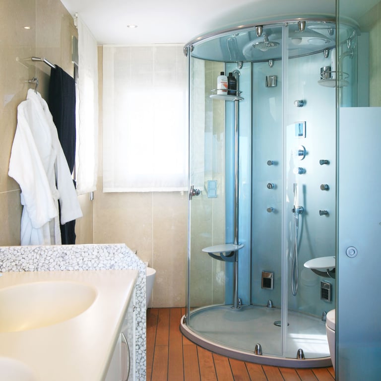 Baño con cabina de ducha de cristal multichorro, encimera de lavabo con textura de guijarros y suelo de madera cálida. La luz natural entra por la ventana.