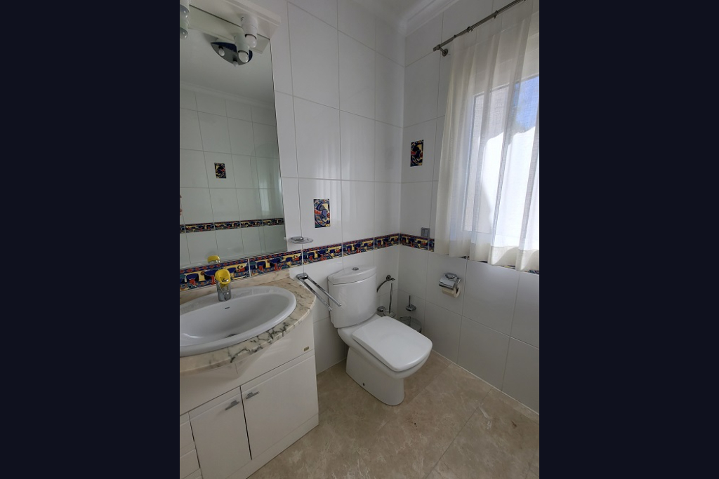 Baño con paredes de azulejos blancos y cenefa decorativa. Presenta un lavabo con encimera de mármol claro, inodoro y ventana con cortina traslúcida para luz natural.