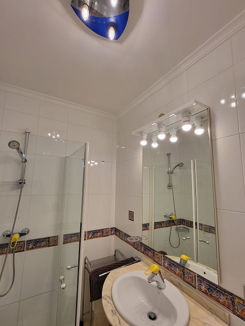 Baño funcional con azulejos blancos y cenefa decorativa, ducha con mampara de cristal y lavabo con encimera efecto mármol. Espejo iluminado y lámpara de techo azul y plata.