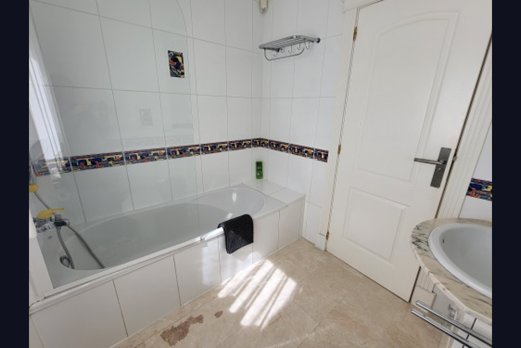 Baño funcional con bañera blanca, mampara de cristal y paredes alicatadas en blanco con cenefa decorativa. Incluye lavabo con encimera de mármol y luz natural.