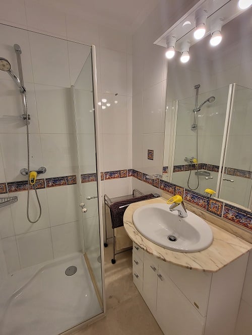 Baño funcional con ducha acristalada, tocador con encimera de mármol claro y espejo iluminado. Azulejos blancos con cenefa decorativa mediterránea.