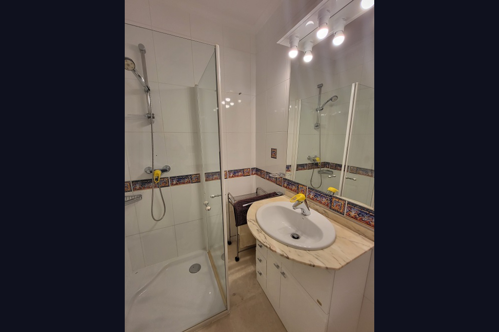 Baño funcional con ducha de mampara de cristal, lavabo sobre encimera de mármol claro y mueble blanco. Las paredes presentan azulejos blancos con un borde decorativo de estilo mediterráneo.