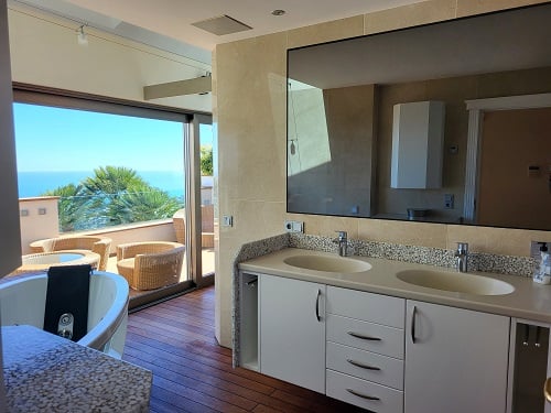 Baño principal con doble lavabo y bañera de hidromasaje, conectado a una terraza de madera con mobiliario de mimbre y vistas directas al mar Mediterráneo.