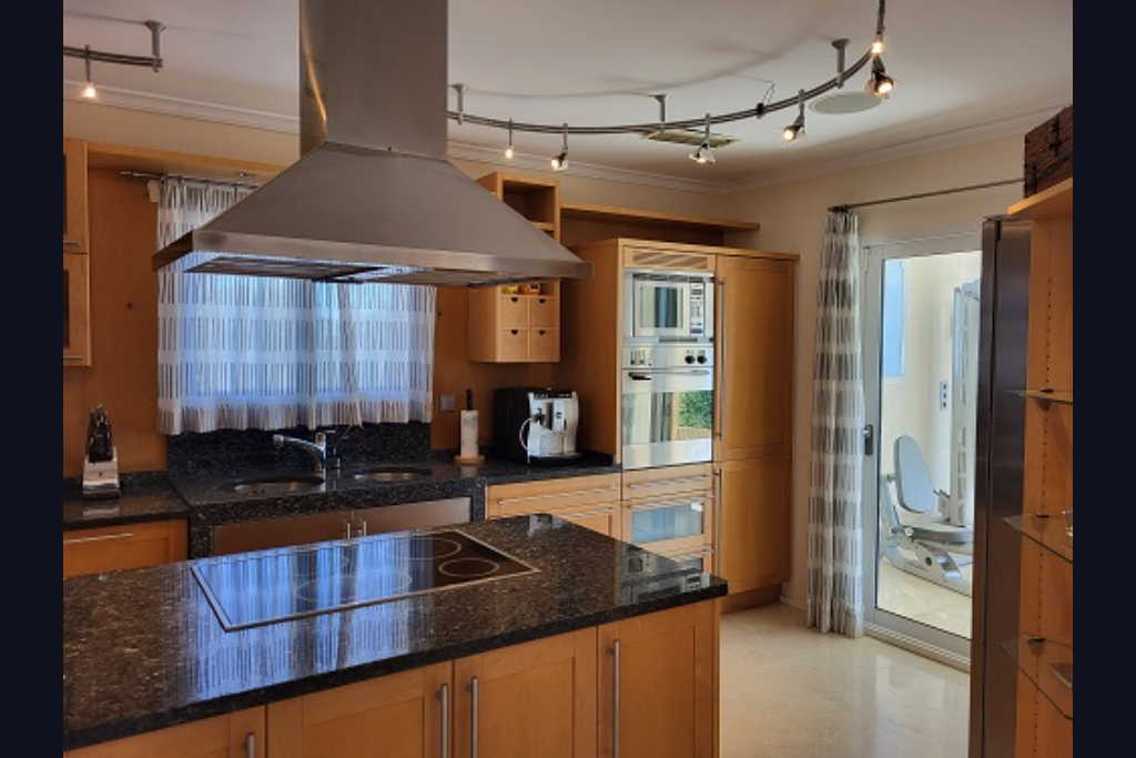 Cocina moderna con armarios de madera clara, encimeras de granito oscuro, isla central con placa de inducción y electrodomésticos de acero inoxidable. Acceso a gimnasio.