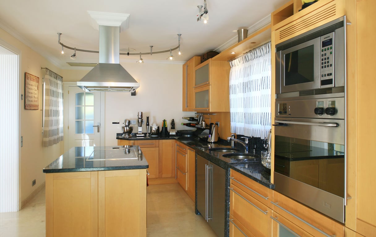Cocina moderna con muebles de madera clara, encimeras de granito oscuro y una isla central con vitrocerámica integrada. Electrodomésticos de acero inoxidable incluyen horno, microondas y campana extractora.