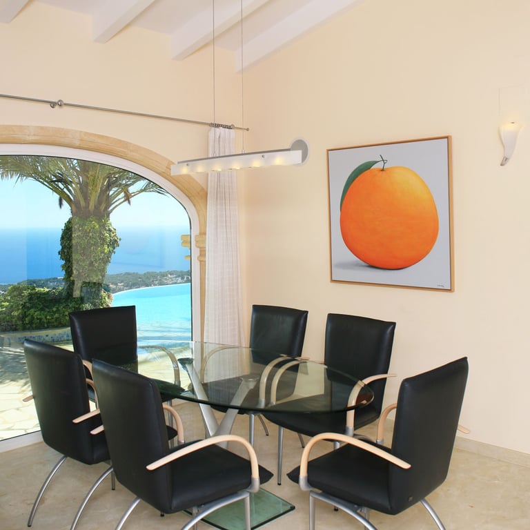Comedor con mesa de cristal y sillas modernas, destacando un gran ventanal arqueado que ofrece vistas panorámicas al mar y la costa. Un cuadro de un naranja decora la pared.