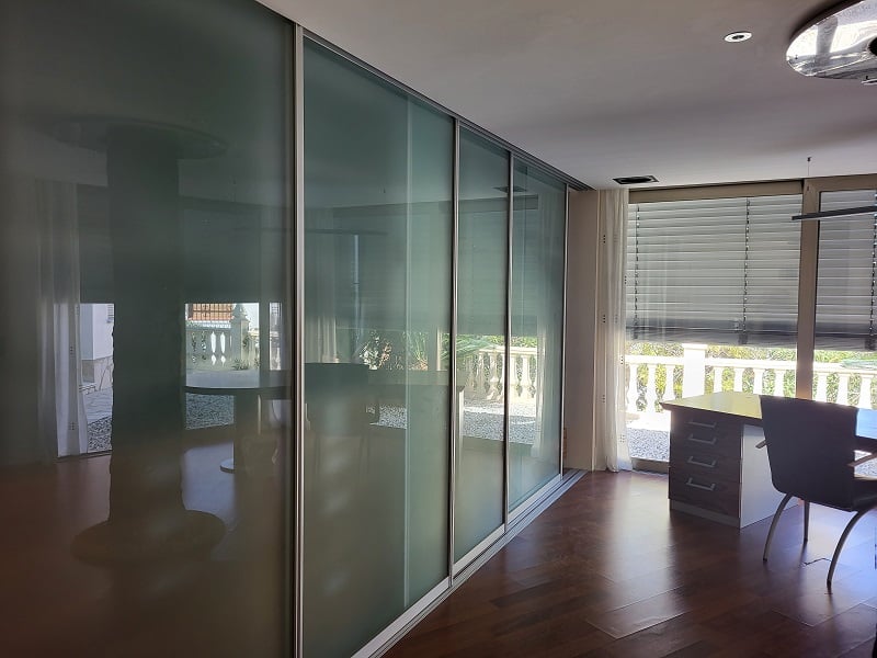 Despacho o dormitorio con elegantes puertas correderas de cristal esmerilado, suelo de madera oscura y zona de trabajo junto a la ventana con vistas a la balaustrada.