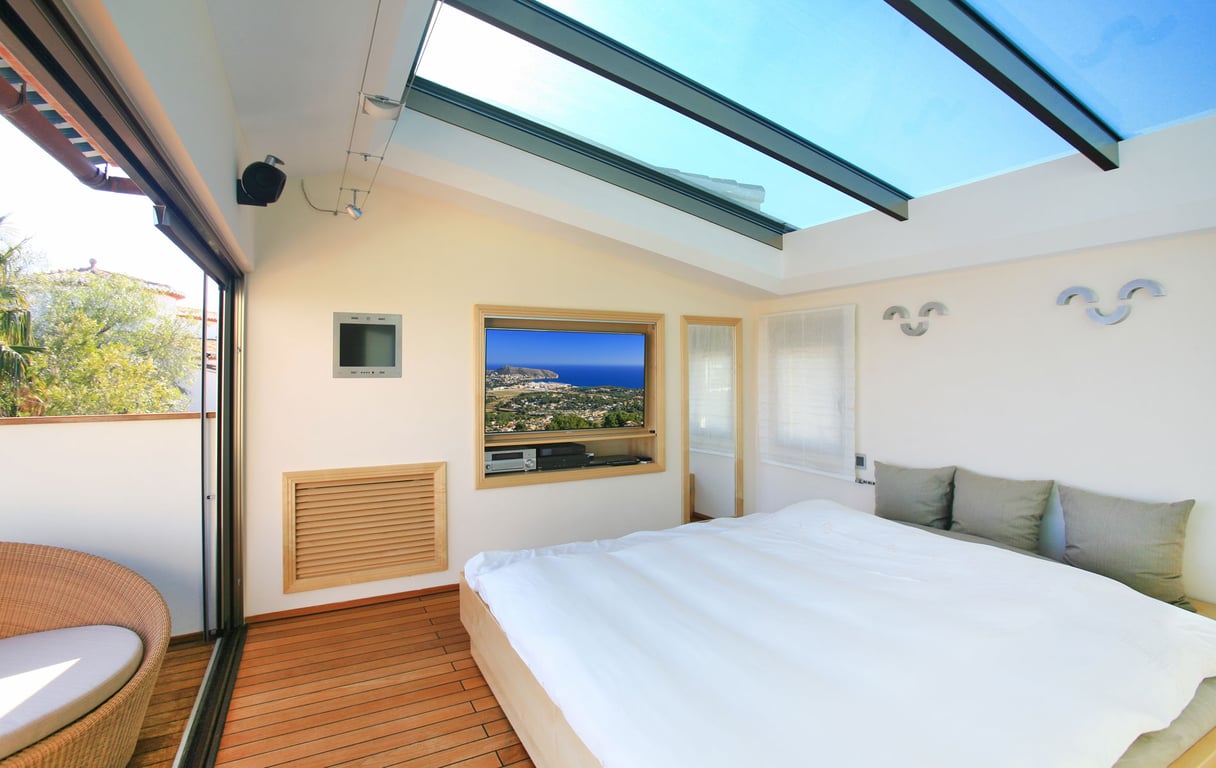 Dormitorio con amplio lucernario de cristal y suelos de madera. Una puerta corredera da acceso a una terraza privada con sillón de mimbre y TV empotrada con vistas costeras.