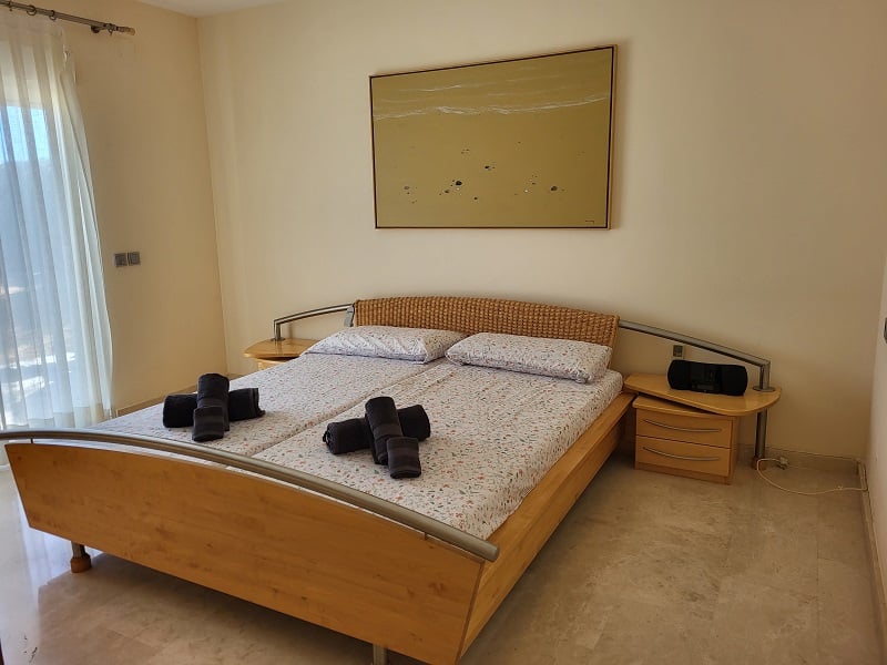 Dormitorio con cama de madera y cabecero de mimbre, flanqueada por mesitas de noche con cajones. Un cuadro abstracto adorna la pared sobre la cama, y la luz natural entra por un ventanal con cortinas claras.