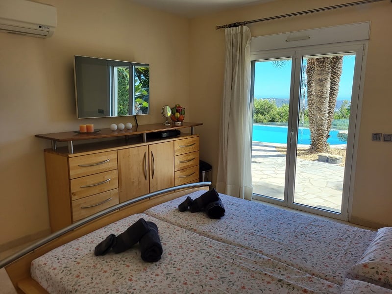 Dormitorio con dos camas individuales, TV de pared sobre cómoda de madera y acceso directo a la terraza exterior con piscina y palmeras.
