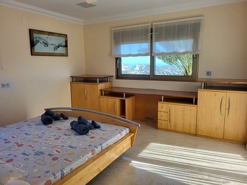 Dormitorio luminoso con cama de madera clara, escritorio integrado y armarios. Las ventanas ofrecen vistas panorámicas a la costa, iluminando la estancia con luz natural.