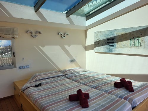 Dormitorio luminoso con dos camas individuales, ropa de cama a rayas y toallas rojas. Destaca un tragaluz que inunda la estancia de luz natural y un cuadro abstracto en la pared.