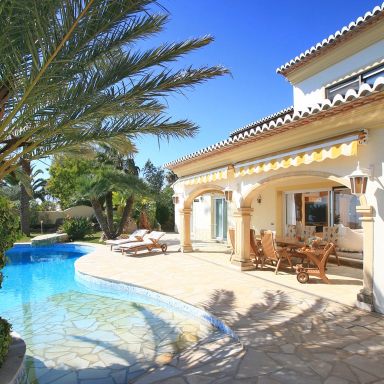 El exterior de la villa presenta una piscina de forma libre y una amplia terraza de piedra que conduce a una naya cubierta con mobiliario de comedor de madera y toldos rayados.