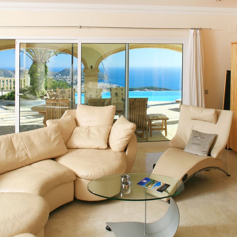 Espaciosa sala de estar con sofás curvos en tonos claros y suelo de mármol, ofreciendo acceso directo a la terraza con piscina infinity y vistas panorámicas al mar Mediterráneo.