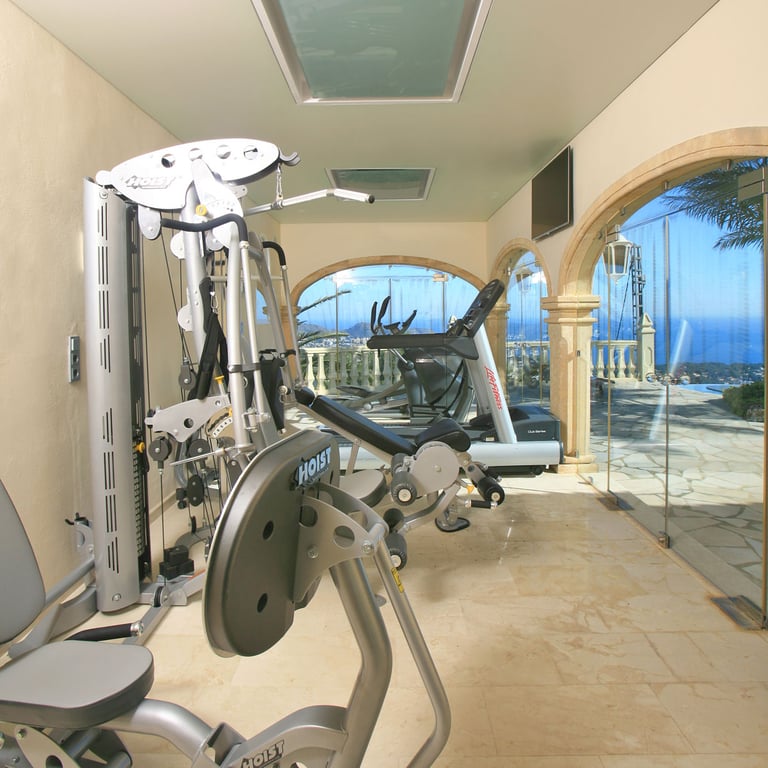 Gimnasio privado con equipo Hoist y Life Fitness, suelos de mármol y ventanales arqueados con vistas panorámicas al mar y la piscina. Claraboyas aportan luz natural.