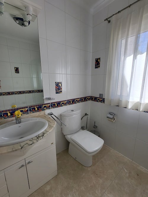Luminoso baño con azulejos blancos, cenefa decorativa colorida y piezas de acento. Cuenta con un tocador de mármol con lavabo integrado, inodoro moderno y ventana con cortina traslúcida.