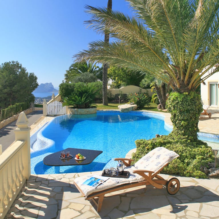 Piscina curva con una mesa negra sumergida y refrescos, junto a una terraza de piedra con tumbona. Se aprecian vistas lejanas al mar y una formación rocosa bajo un cielo despejado.