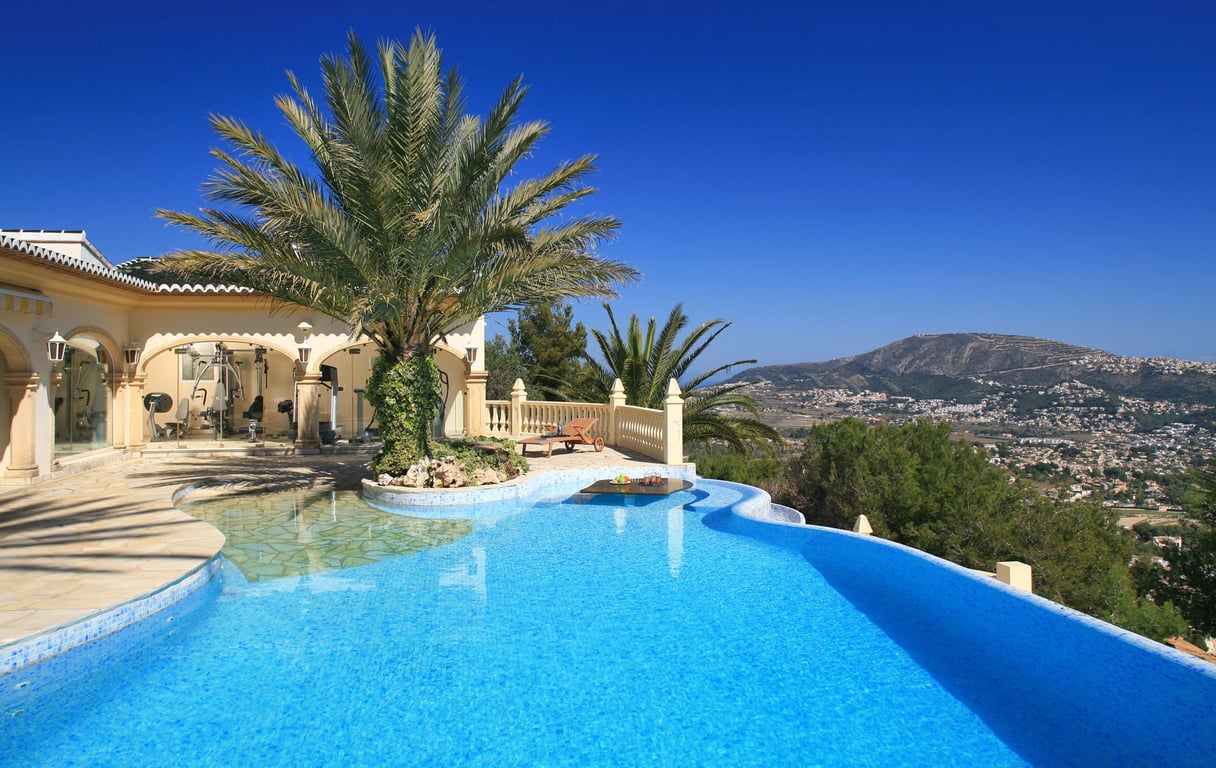 Piscina curva de mosaico azul con isla de palmeras y mesa sumergida. Gimnasio acristalado y terraza con vistas panorámicas a la montaña.