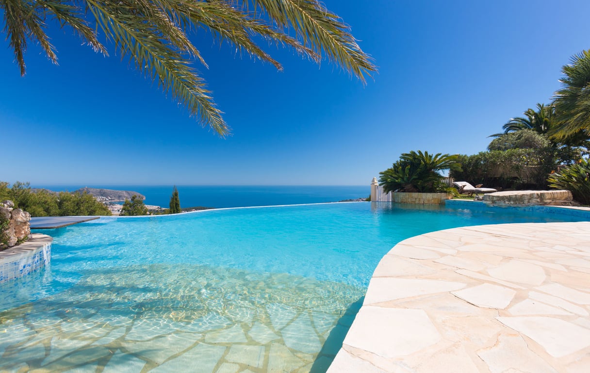 Piscina infinity con entrada tipo playa y bordes de piedra clara, ofreciendo vistas panorámicas al Mediterráneo y la costa, bajo un cielo azul.