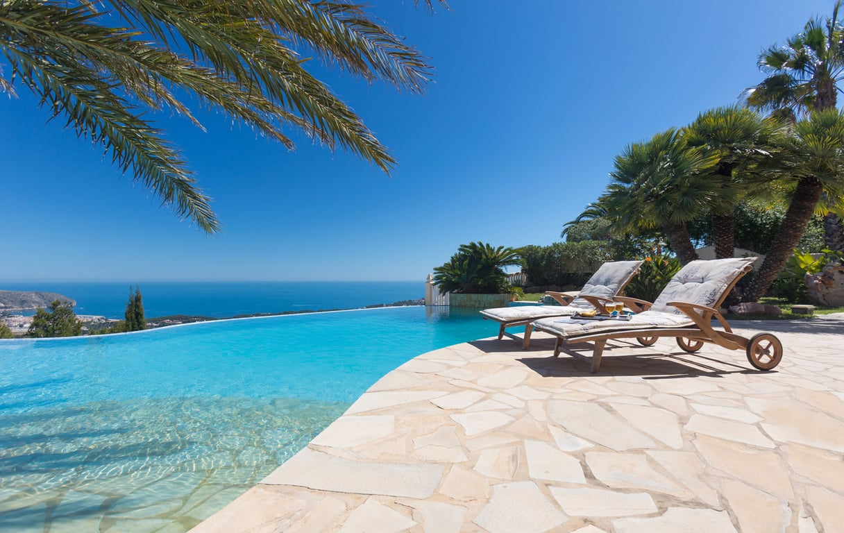 Terraza con piscina infinita y dos tumbonas de madera sobre un patio de losas, con amplias vistas al mar Mediterráneo y la costa lejana.