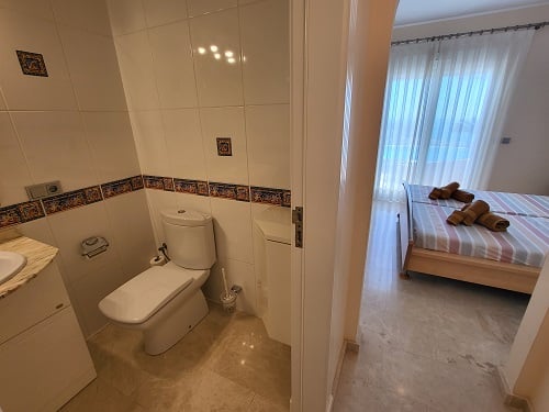 Vista desde un baño alicatado con inodoro y lavabo, hacia un luminoso dormitorio con camas individuales, ropa de cama a rayas y gran ventanal con cortinas.