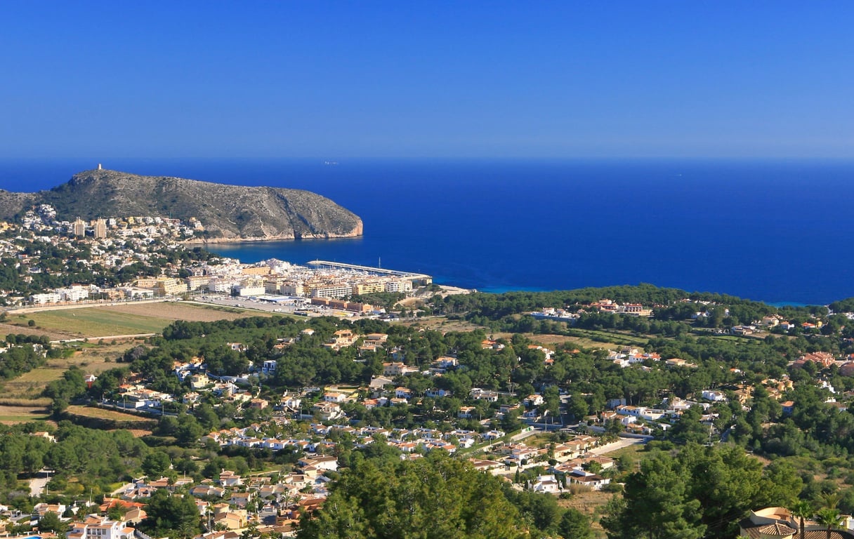 Vista panorámica desde una colina, mostrando un pueblo costero, un puerto deportivo, una montaña y el mar Mediterráneo bajo un cielo despejado.