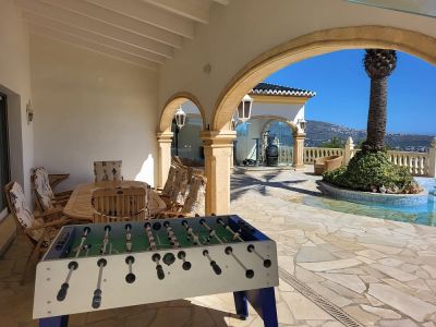 Amplia naya cubierta con mesa de comedor de madera y futbolín, enmarcada por arcos de piedra y un suelo de losas irregulares. Ofrece vistas a la piscina con isla de palmeras y a las montañas lejanas.