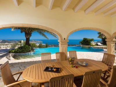 Amplia naya cubierta con mesa de comedor de madera y sillas, ofreciendo vistas panorámicas al mar Mediterráneo y la costa a través de elegantes arcos de piedra y una piscina infinity con palmeras.