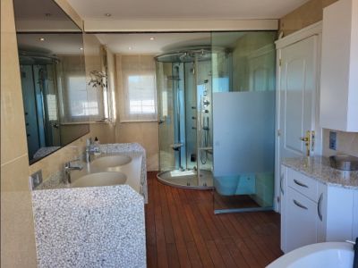 Amplio baño con doble lavabo de diseño en guijarro, ducha circular de hidromasaje y suelos de madera. Incluye espejo grande y ventanas con persianas.