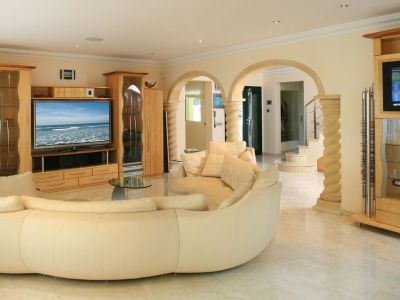 Amplio salón con sofá curvo color crema, suelos de mármol claro y muebles de entretenimiento de madera clara a medida. Arcos de piedra con columnas decoradas conectan con la entrada.