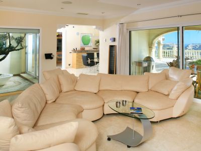 Amplio salón con sofá seccional claro, mesa de centro de cristal y suelos de mármol. Grandes ventanales conectan con una terraza con vistas al mar y un patio interior con olivo.