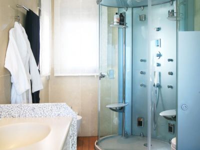 Baño con cabina de ducha de cristal multichorro, encimera de lavabo con textura de guijarros y suelo de madera cálida. La luz natural entra por la ventana.