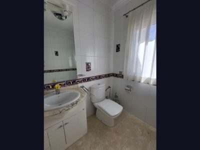 Baño con paredes de azulejos blancos y cenefa decorativa. Presenta un lavabo con encimera de mármol claro, inodoro y ventana con cortina traslúcida para luz natural.
