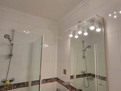 Baño funcional con azulejos blancos y cenefa decorativa, ducha con mampara de cristal y lavabo con encimera efecto mármol. Espejo iluminado y lámpara de techo azul y plata.