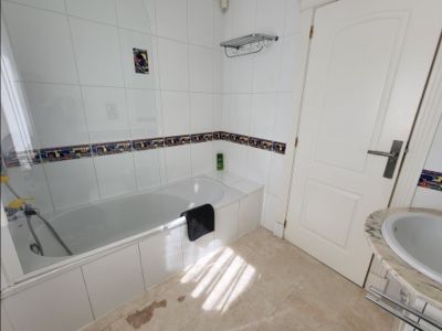 Baño funcional con bañera blanca, mampara de cristal y paredes alicatadas en blanco con cenefa decorativa. Incluye lavabo con encimera de mármol y luz natural.