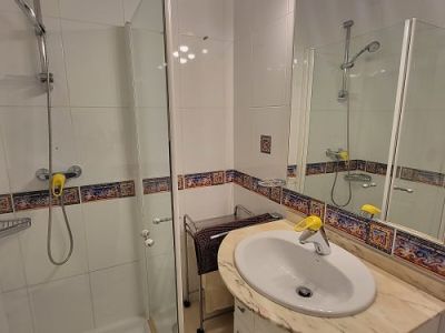 Baño funcional con ducha acristalada, tocador con encimera de mármol claro y espejo iluminado. Azulejos blancos con cenefa decorativa mediterránea.