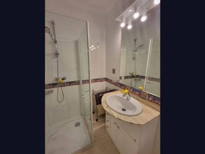 Baño funcional con ducha de mampara de cristal, lavabo sobre encimera de mármol claro y mueble blanco. Las paredes presentan azulejos blancos con un borde decorativo de estilo mediterráneo.