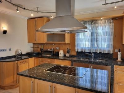 Cocina funcional con armarios de madera clara y encimeras de granito oscuro. La isla central integra una placa de inducción bajo una campana extractora de acero inoxidable.