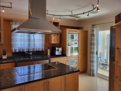 Cocina moderna con armarios de madera clara, encimeras de granito oscuro, isla central con placa de inducción y electrodomésticos de acero inoxidable. Acceso a gimnasio.