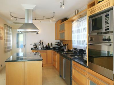 Cocina moderna con muebles de madera clara, encimeras de granito oscuro y una isla central con vitrocerámica integrada. Electrodomésticos de acero inoxidable incluyen horno, microondas y campana extractora.