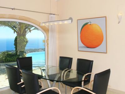 Comedor con mesa de cristal y sillas modernas, destacando un gran ventanal arqueado que ofrece vistas panorámicas al mar y la costa. Un cuadro de un naranja decora la pared.
