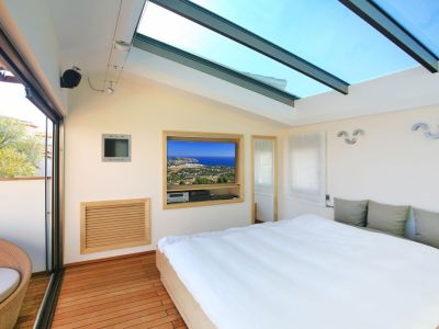 Dormitorio con amplio lucernario de cristal y suelos de madera. Una puerta corredera da acceso a una terraza privada con sillón de mimbre y TV empotrada con vistas costeras.